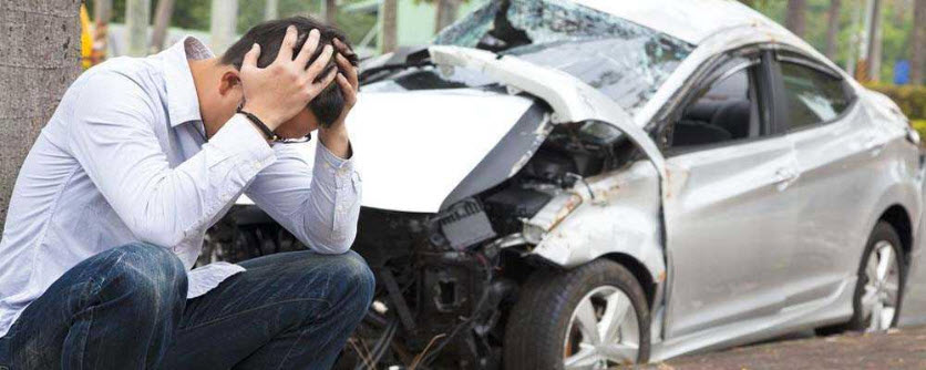 Man sitting on curb holding his head with crushed car behind him